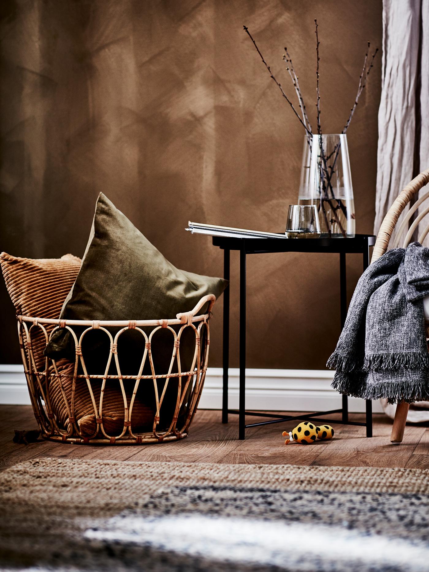 A rustic room with a SNIDAD rattan basket with warm-toned cushions in it next to a black tray table and a rattan armchair.