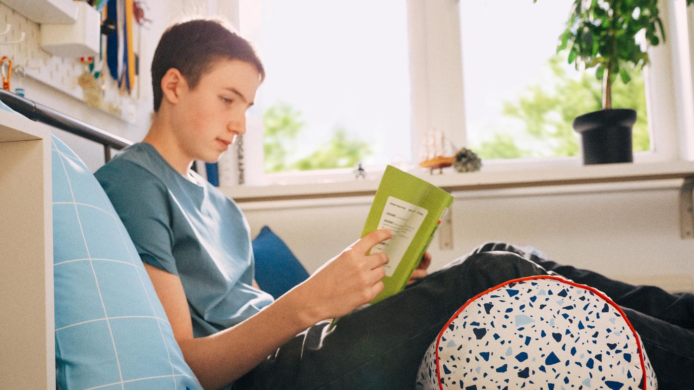 A schoolchild sits reading on his bed, leaning back against a pillow with his legs supported by a MÖJLIGHET cushion.