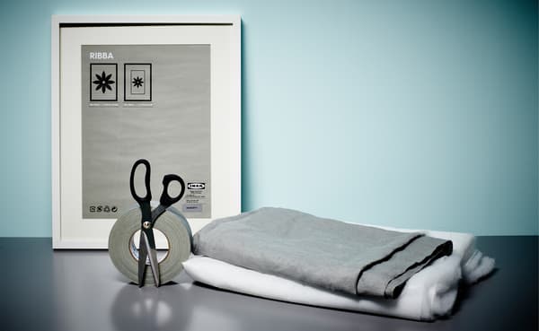 A scissor, tape, a photo frame, grey fabric and white wadding on a table. They will be used to make a soundproofed wall.