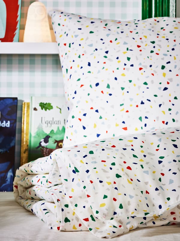 A set of MÖJLIGHET bed linen on an unmade bed, with a SOLBO LED lamp and books on picture ledges on a wall behind.