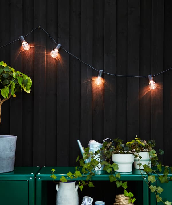 A shimmering lighting chain hanging from a dark brown house wall, lighting up the patio and two green cabinets.