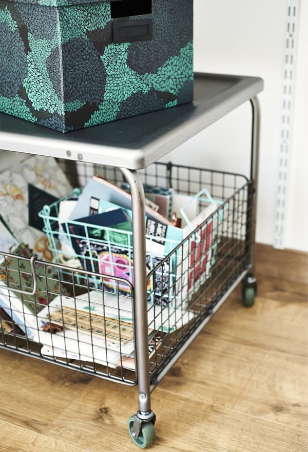 A short stainless steel trolley with green rubber castors and a wire basket to store stationary.
