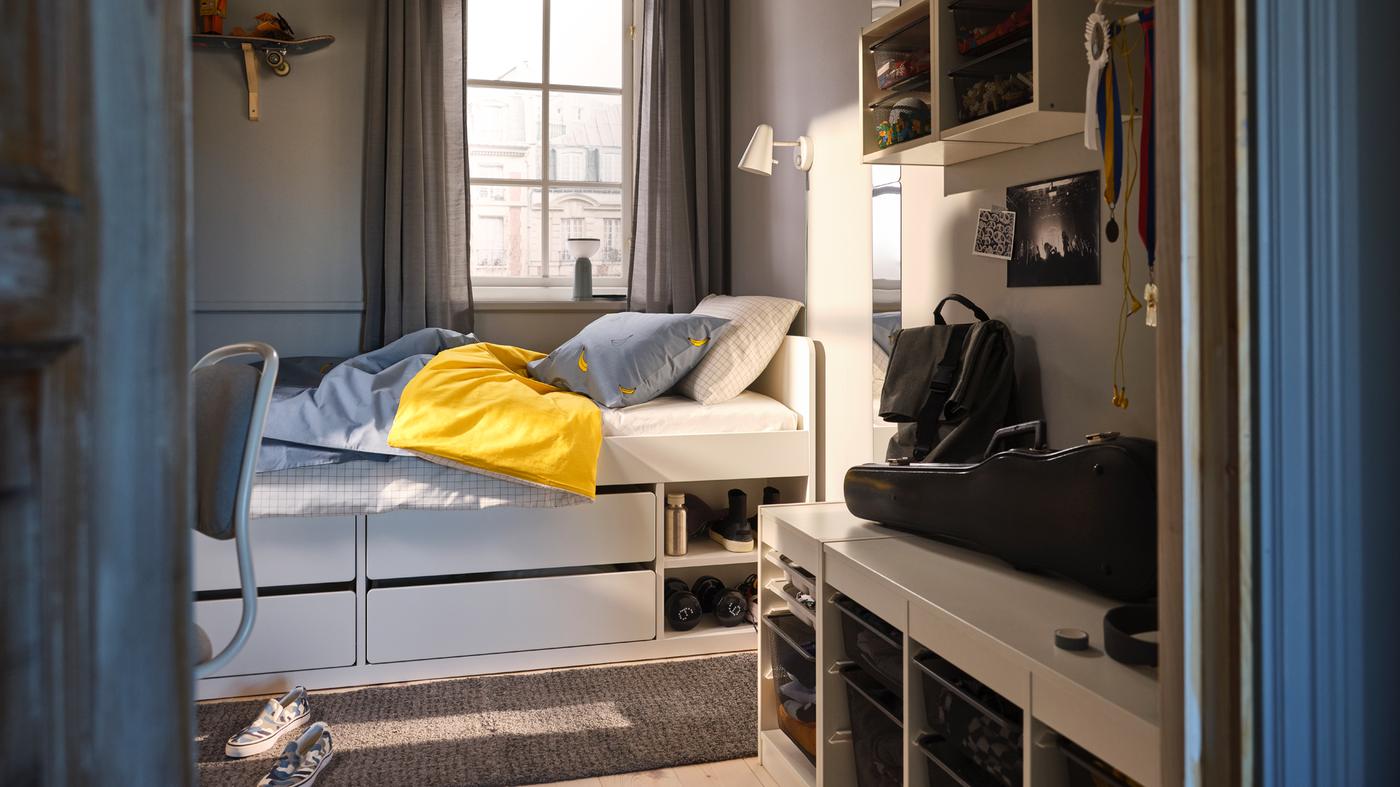 A SLÄKT bed with in blue bed linen in front of a window in a small bedroom with TROFAST storage units on the wall and floor.
