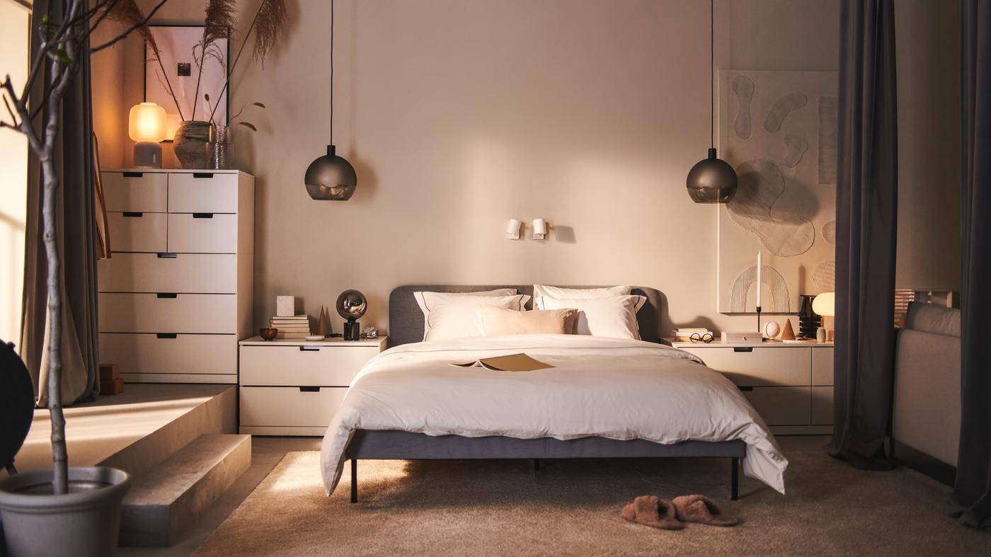 A SLATTUM upholstered bed and various white NORDLI chests of drawers stand against a wall in a bedroom.
