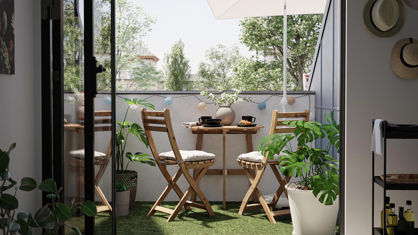 A small balcony with wooden tables and chairs, artificial grass decking, and a Monstera plant in a white plant pot.