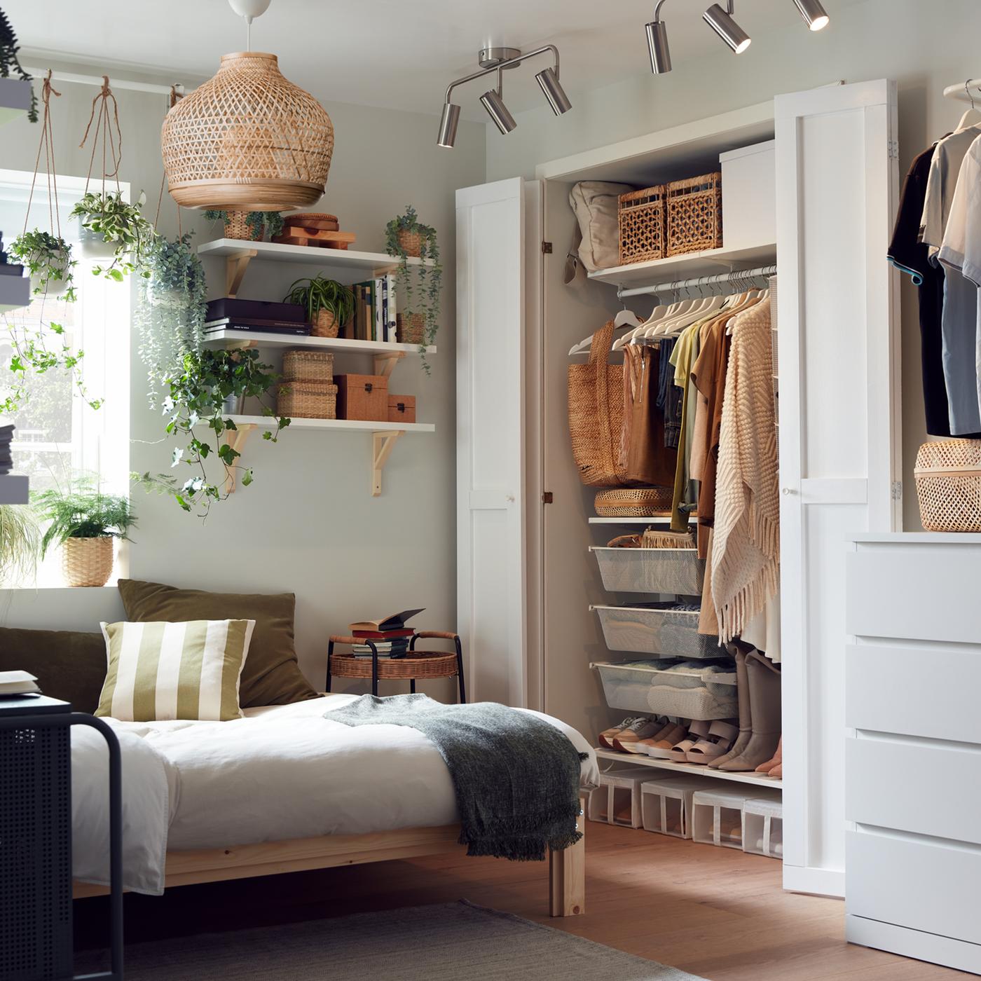 A small bedroom with a wooden bed frame, an opened wardrobe with neatly organised clothes, shelves with boxes and plants.