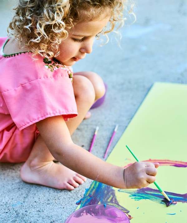 A small child painting bright colours on yellow paper, on a concrete floor.