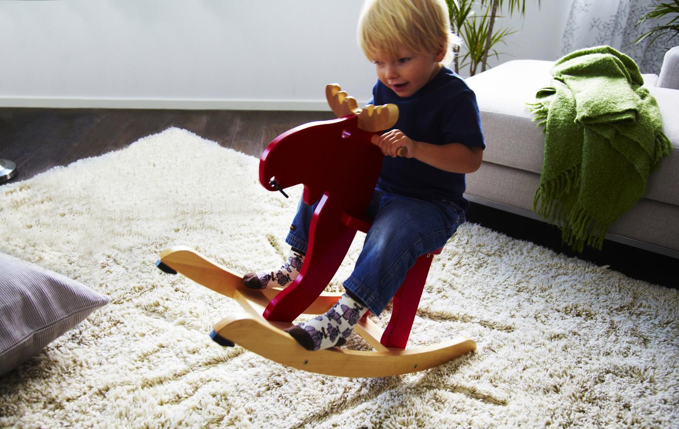 A small child playing on a red moose-shaped rocking horse in a living room with neutral decor.