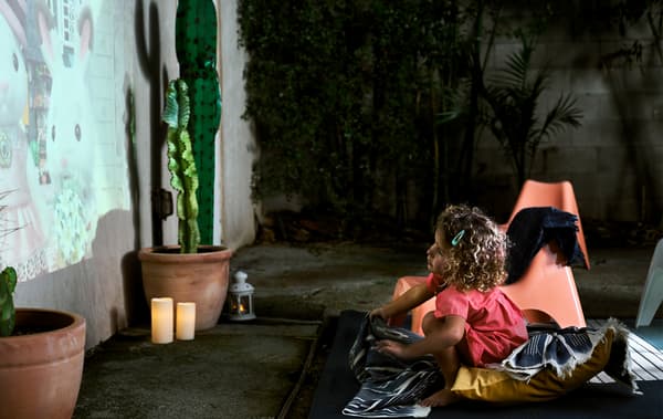 A small child sitting on blankets and cushions watching a cartoon projected on an exterior wall, next to a potted cactus and LED candles.
