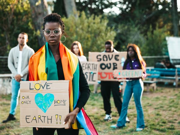 A small group of climate activists are holding handwritten, brown cardboard signs in peaceful protest in a park setting.