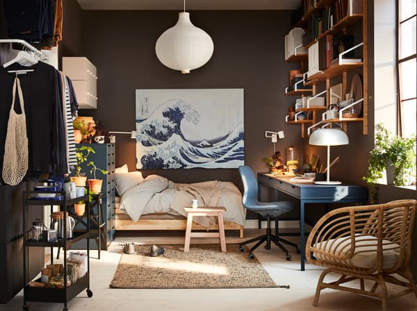 A small home with a dark blue desk, a rattan armchair, a white pendant lamp shade and a painting with a big blue wave.