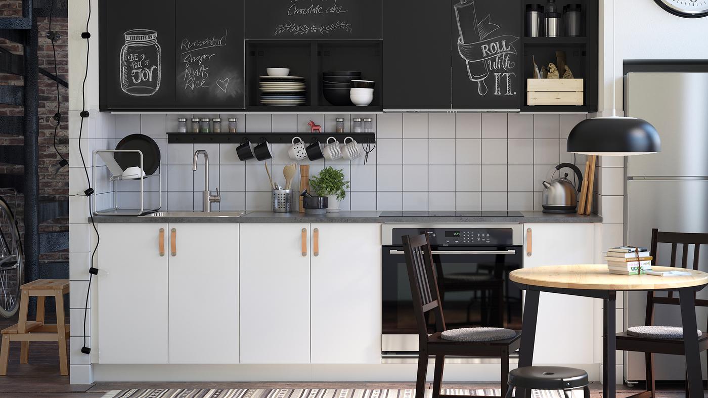 A small kitchen with white cabinets at the bottom and chalk doors inscribed with messages above, a table and two chairs.