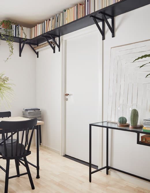 A small room with a dining table, glass desk and black book shelves mounted high along the ceiling to make it feel airy.