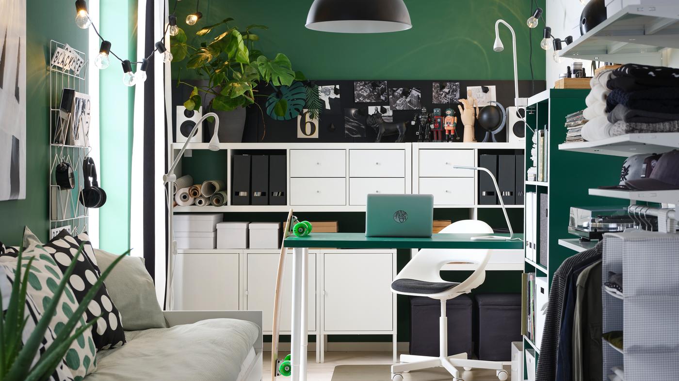 A small room with a green table, white shelving units, a day-bed, an open wardrobe and a black pendant lamp.