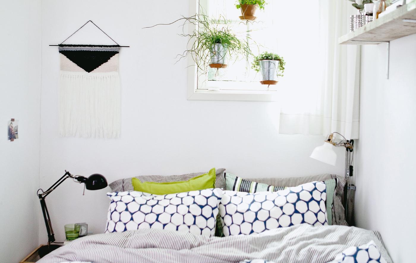 A small room with double bed, hanging plants and shelf.