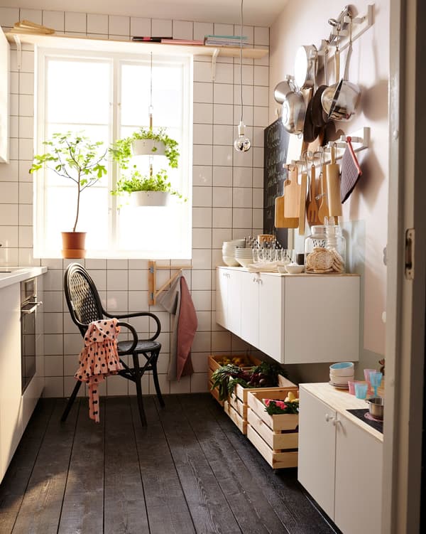 A small, white kitchen with light streaming in the window. There’s a single black chair and a wall of cooking utensils.