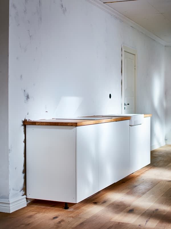 A small, white, straight-file kitchen with a wooden worktop in an otherwise empty room with wooden floor and white walls.