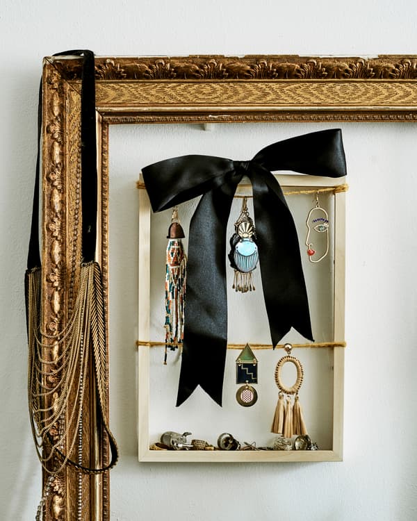 A small wood frame used to display dangly earrings and a black bow, hangs inside a large gilt frame.