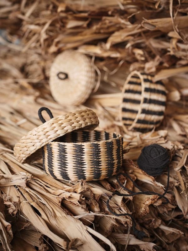 A small woven MÄVINN box with a lid, featuring a black stripe pattern, placed on a pile of natural banana fibre.