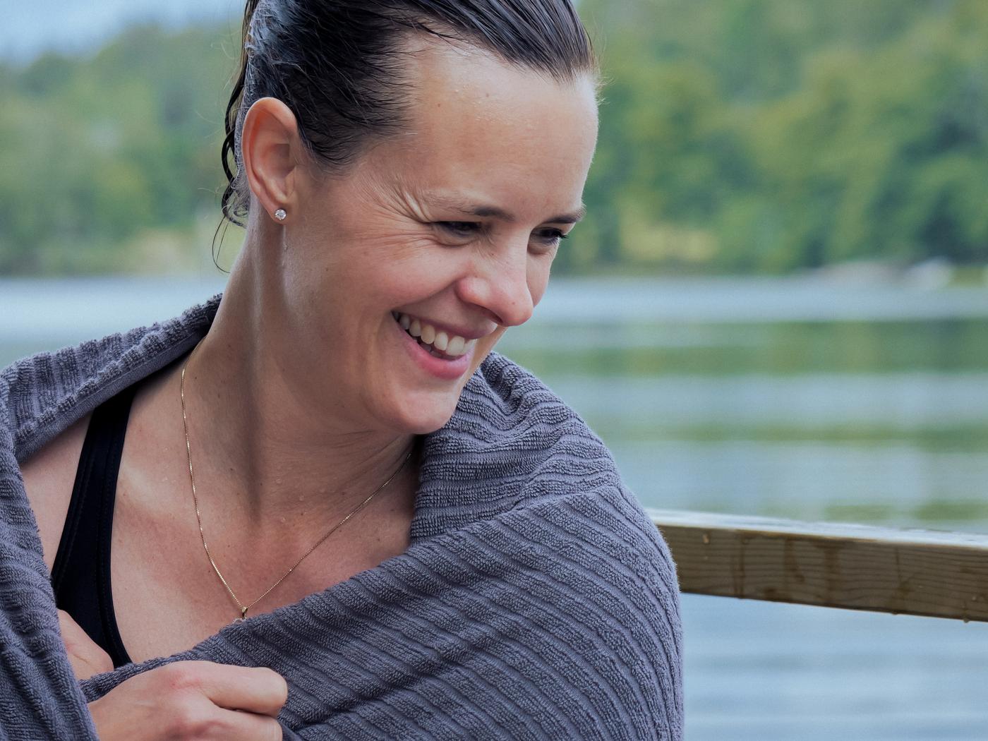 A smiling woman, wrapped in a VÅGSJÖN towel after a swim in a lake.