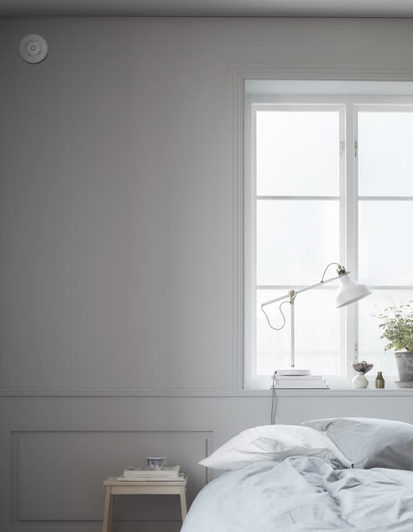 A smoke detector sits high up on a wall near a bed and a step stool.