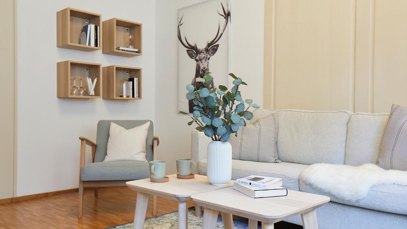 A sofa, armchairs and coffee table in light colours in a living room. Four cube-shaped shelves made of wood hang on the wall.