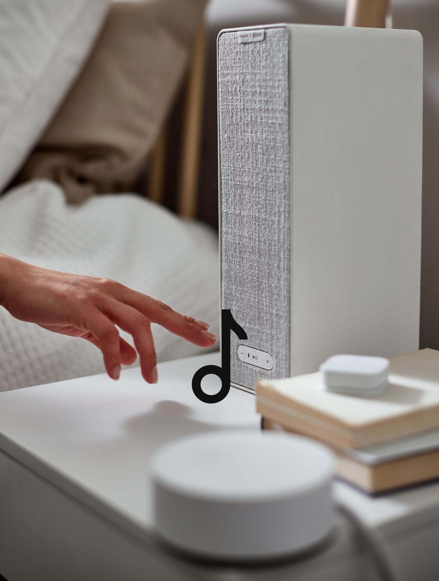 A speaker on a nightstand and a hand reaching to turn it on. A music note icon is on top of the image.