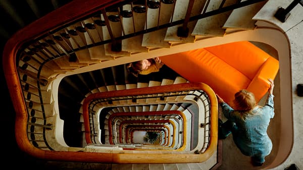 A spiral staircase in a building with many floors, and a young couple carrying a bright orange sofa.