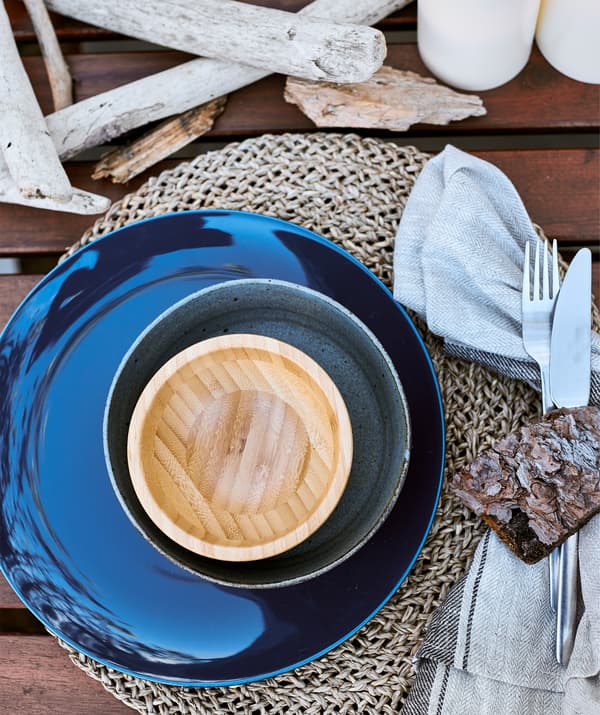 A stack of dark blue and wooden bowls on a woven placemat and driftwood on a wooden table.