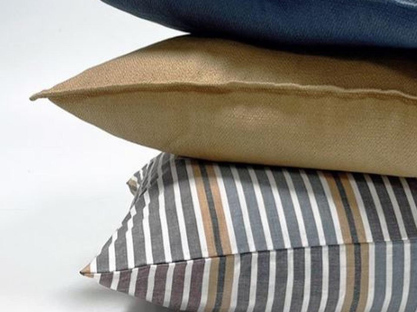 A stack of four decorative pillows with different designs on a light background, including a striped pillow, a golden pillow, a blue pillow, and an off-white knitted throw blanket on top.