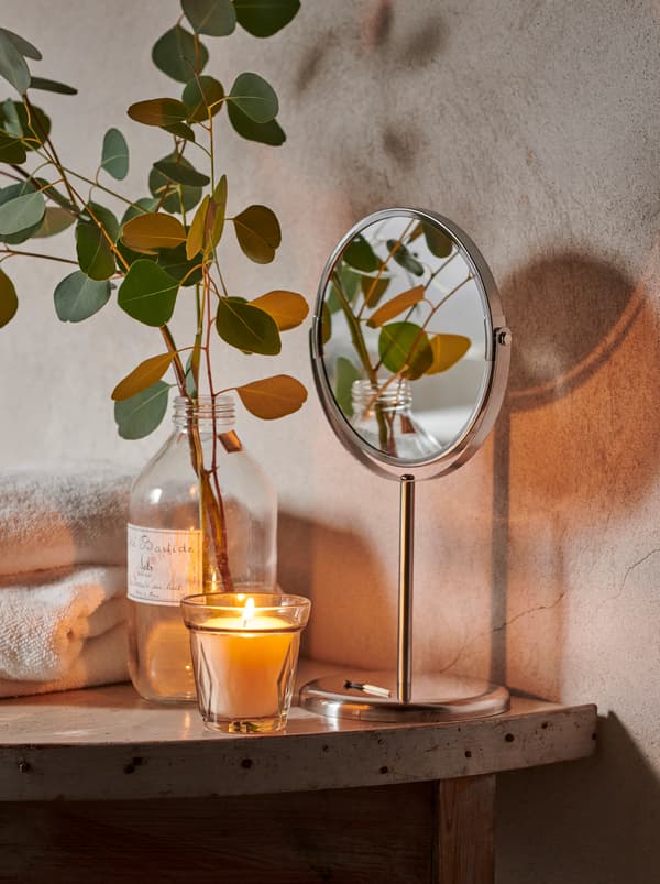 A stack of towels, a VÄLDOFT candle, and a glass bottle with a sprig of eucalyptus leaves on a shelf in a rustic bathroom.