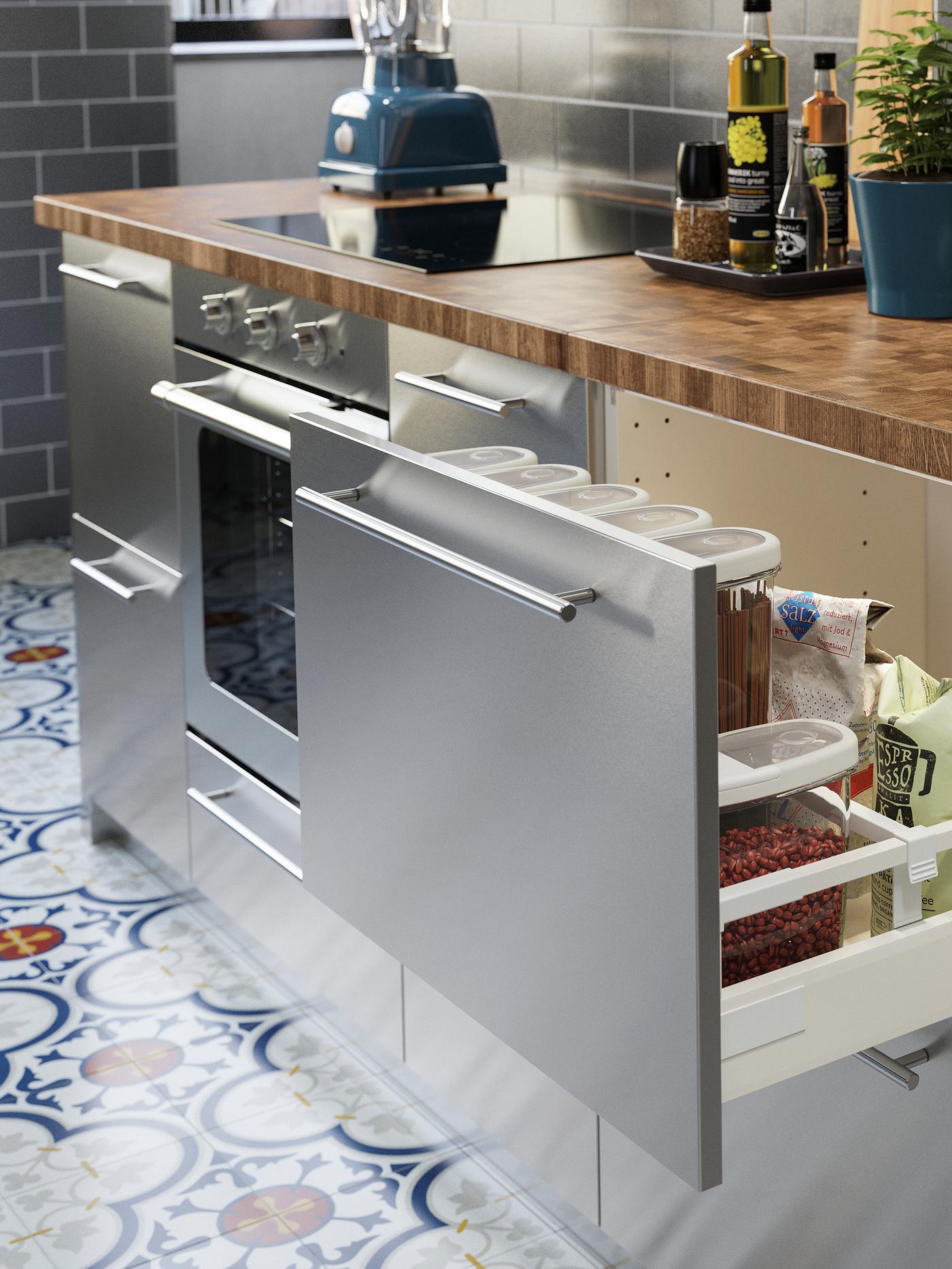 A stainless steel kitchen with a wooden worktop in oak/veneer. One drawer is open, dry goods are stored inside.