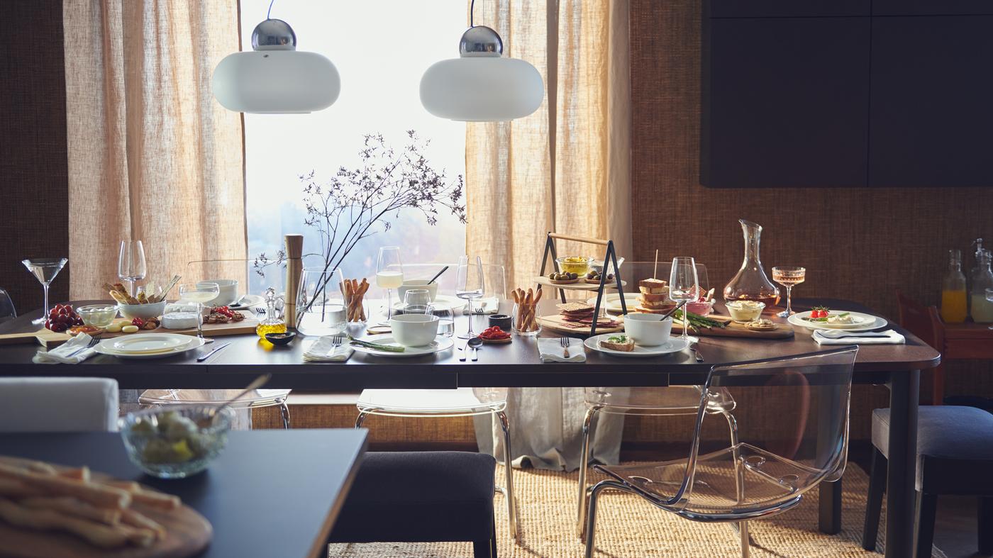 A STRANDTORP extendable dining table in front of a window, laid for a big meal with plates, glasses, flowers and food.
