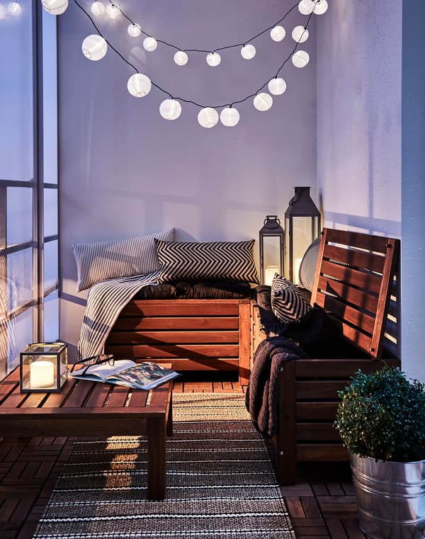 A strings of lit bulbs hang above a cosy balcony corner with soft seating and a coffee table.