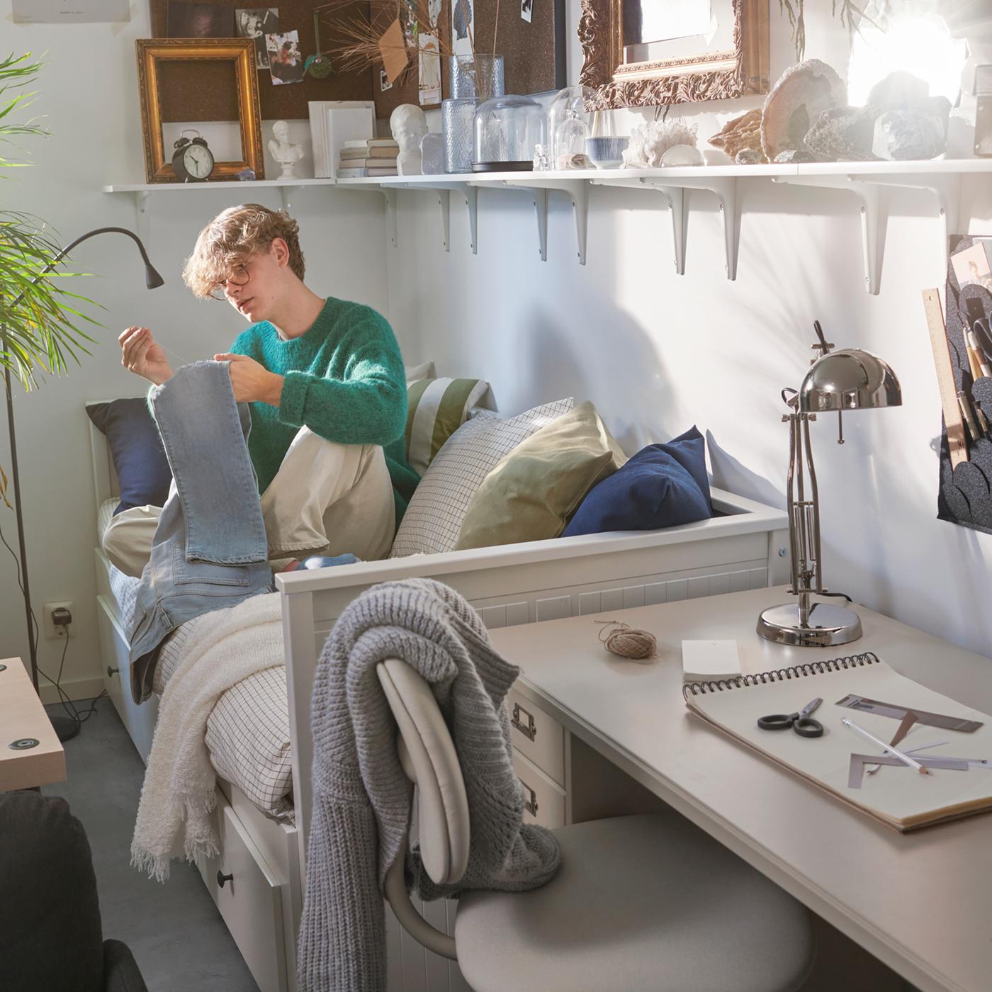 A student sits on a HEMNES day bed in a dorm room. Several BURHULT/SIBBHULT shelves with frames and books are above him.