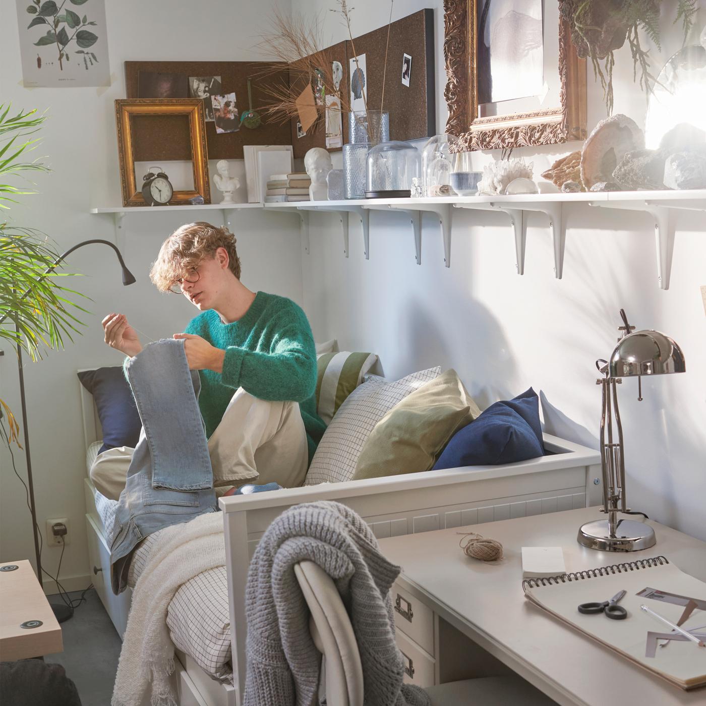 A student sits on a HEMNES day bed sewing a pair of jeans. Shelves on the wall hold an alarm clock, frames and objects.