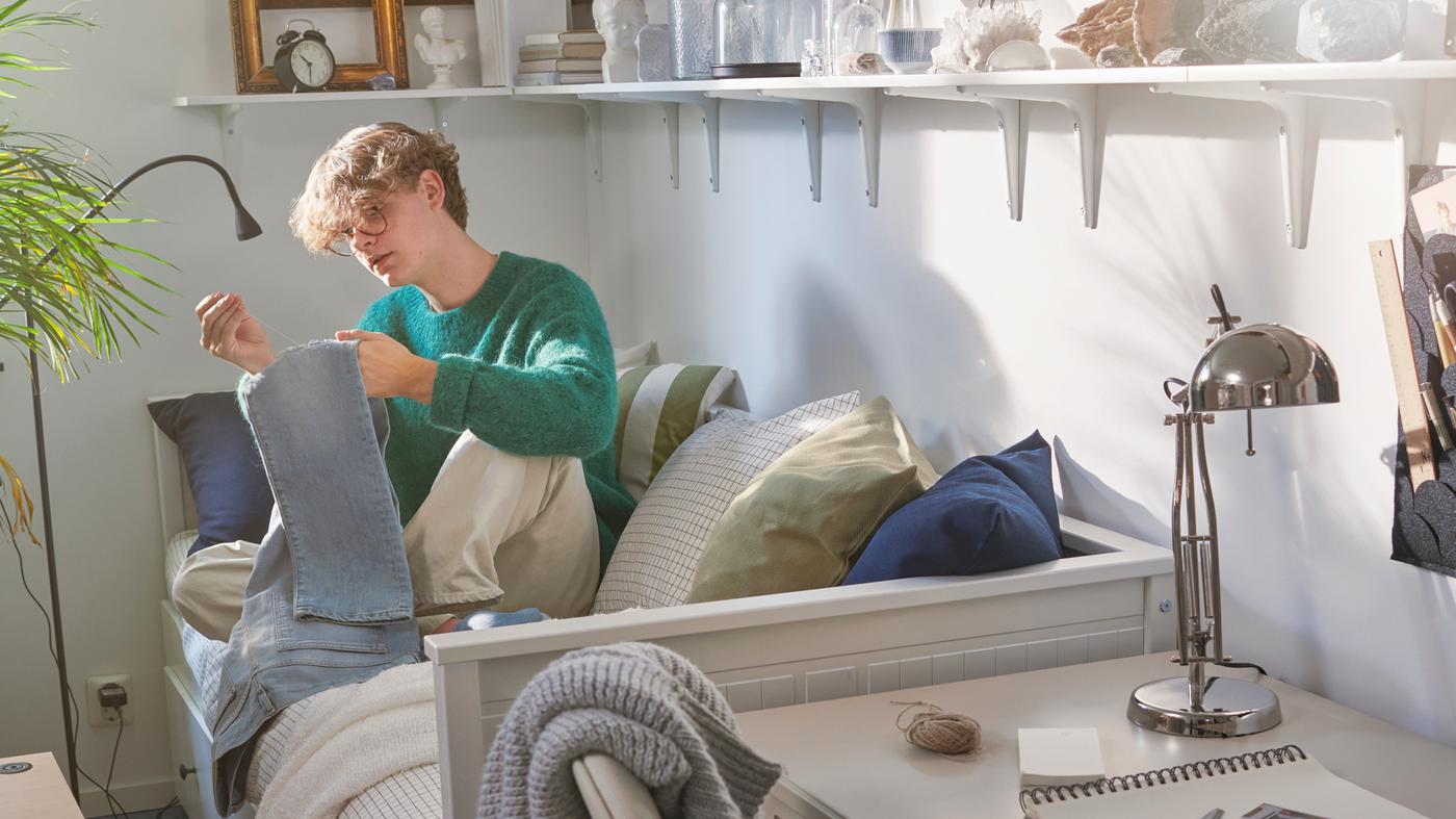A student sits on a HEMNES day bed sewing a pair of jeans. Shelves on the wall hold an alarm clock, frames and objects.