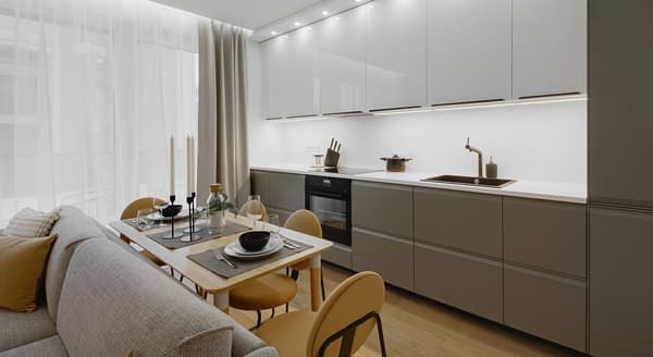 A stylish open-concept kitchen and dining area with sleek white upper cabinets and matte gray lower cabinets, illuminated by recessed ceiling lights and under-cabinet lighting. A wooden dining table is set for four with neutral-toned tableware and black candle holders, adjacent to a light gray sofa with mustard accent pillows, all framed by tall curtains and sheer drapes letting in natural light.