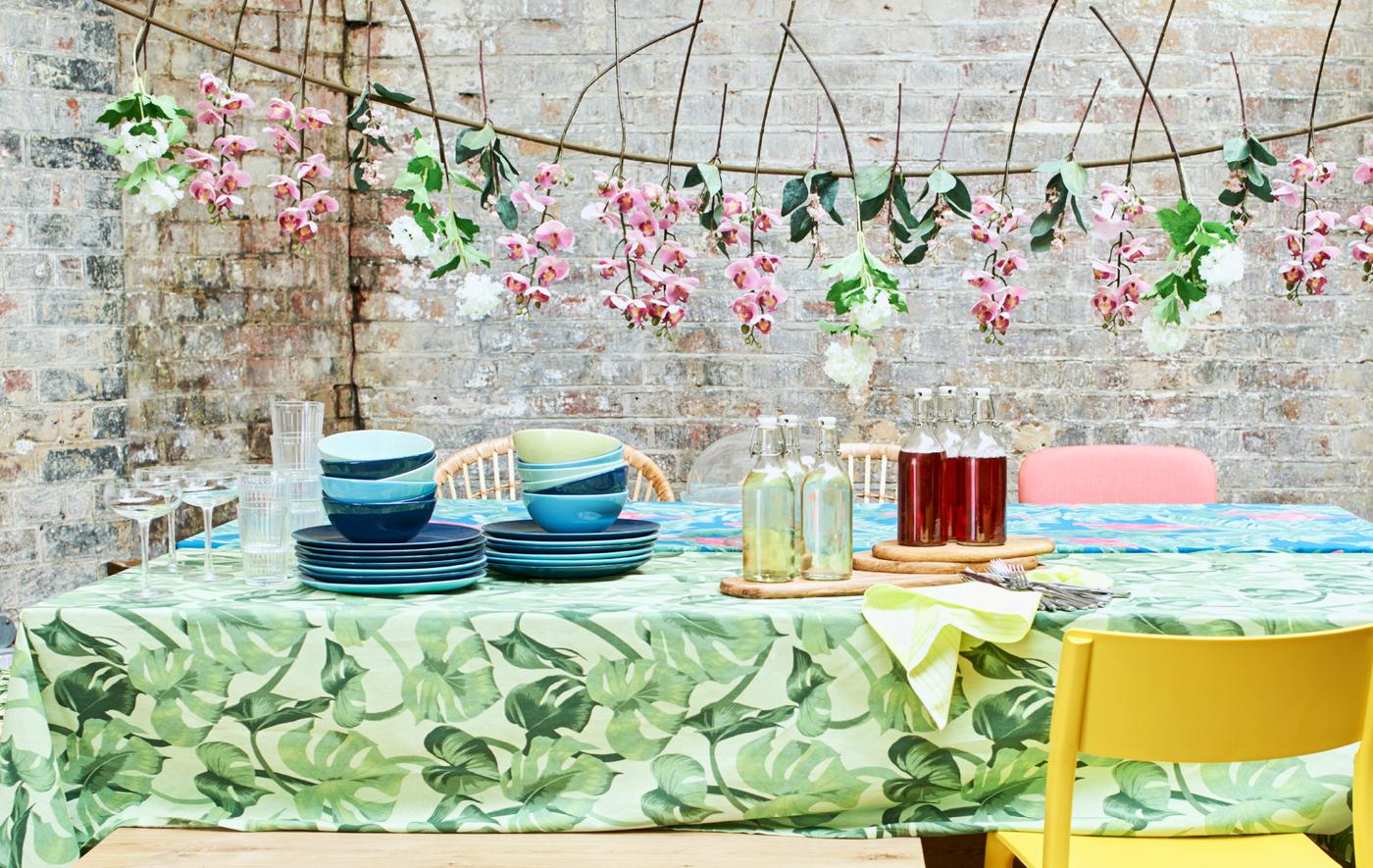 A summer party table with bright tablecloths, piles of blue crockery, mixed chairs and a garland of artificial flowers.