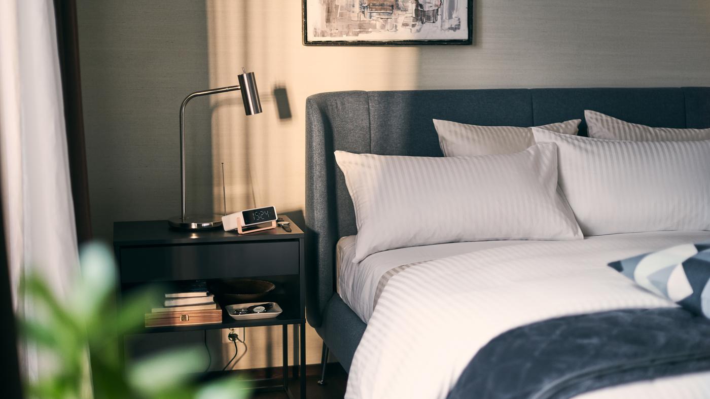 A sunny bedroom with a black bedside table holding books and a lamp beside a blue TUFJORD bed covered in white bed linen.