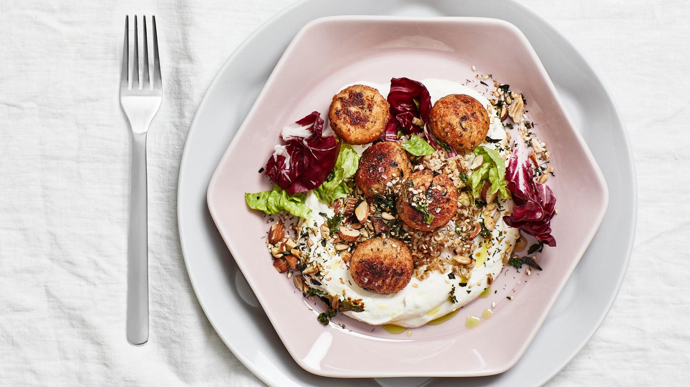 A table set with a white cloth, a fork and a dusty pink octagon shaped plate with five cod balls, greens, nuts and yoghurt.