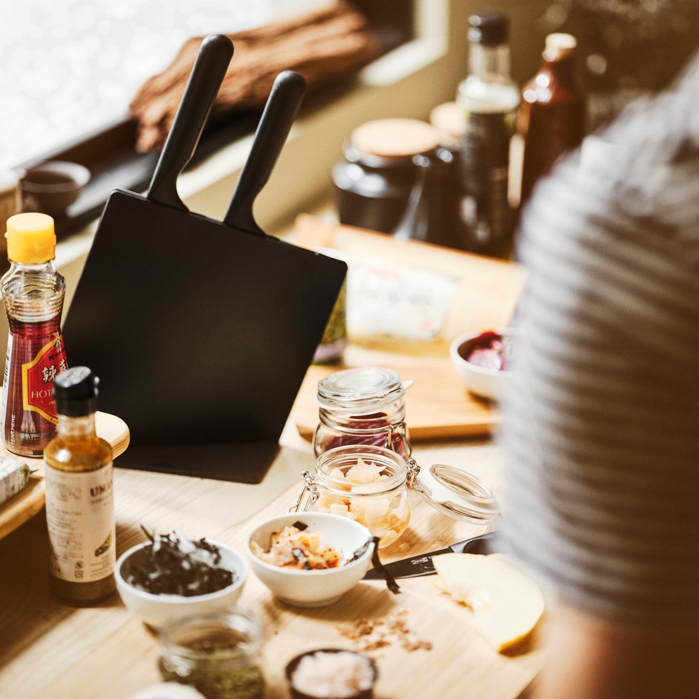 A table surface with a black JÄMFÖRA knife block with knives, an APTITLIG chopping board, KORKEN jars, and white bowls.