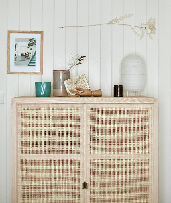 A tall wooden storage cabinet with woven doors set against a panelled wall with a paper lantern and objects displayed on top.