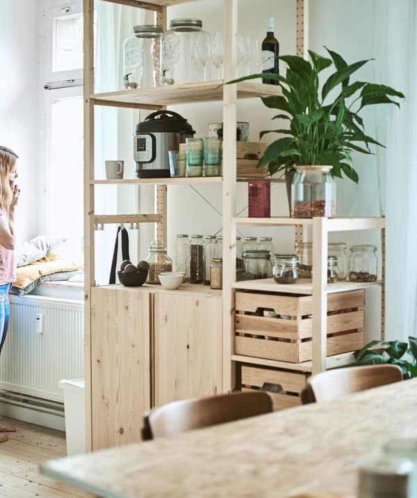 A tall wooden storage unit in a kitchen/diner with glass jars, wood crates and kitchen gadgets on the shelves.