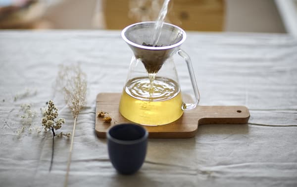 A tea strainer over a glass pot, on a wooden chopping board.