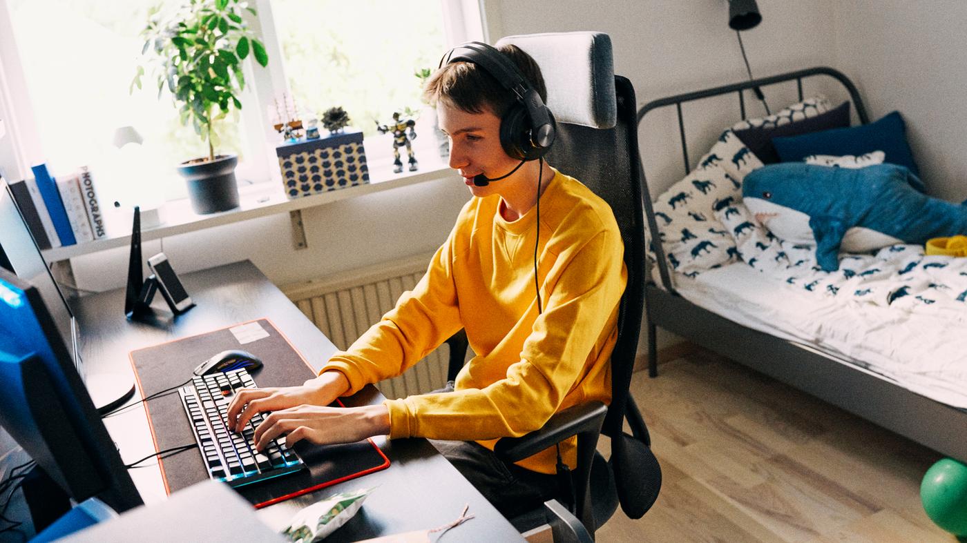 A teen boy in his bedroom sitting on a JÄRVFJÄLLET chair at a LINNMON/ALEX table, using a computer while wearing a headset.