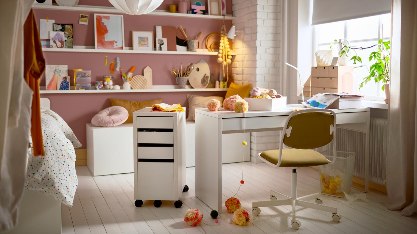 A teenager’s room with a desk, a drawer unit, picture ledges with artsy decorations, cushions, and a swivel chair.