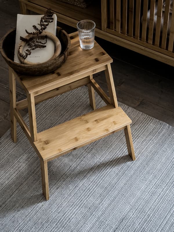 A TENHULT bamboo step stool, on it a glass of water and a small basket, stands on a rug in a wooden-floor bedroom.