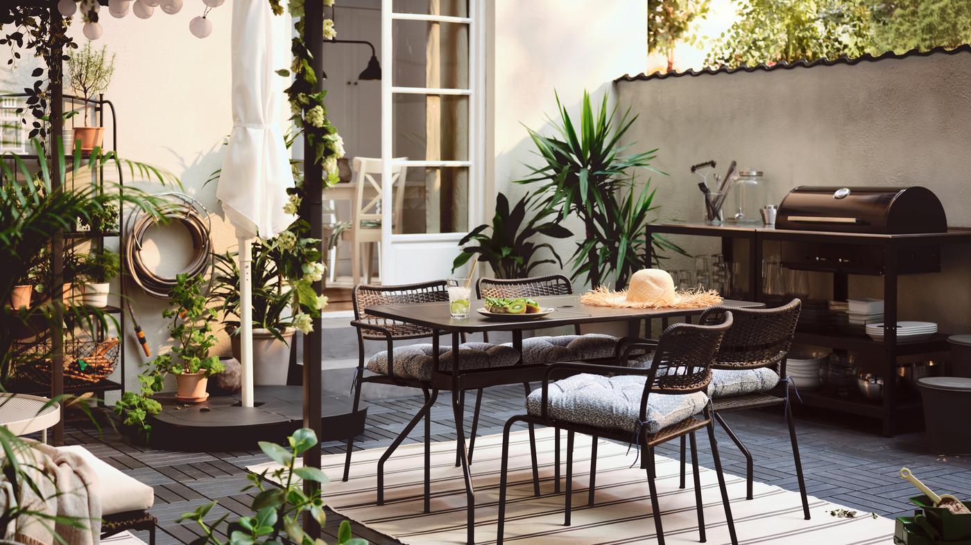A terrace with a VIHOLMEN table surrounded by four LÄCKÖ chairs. An outdoor kitchen with charcoal barbecue is by the wall.