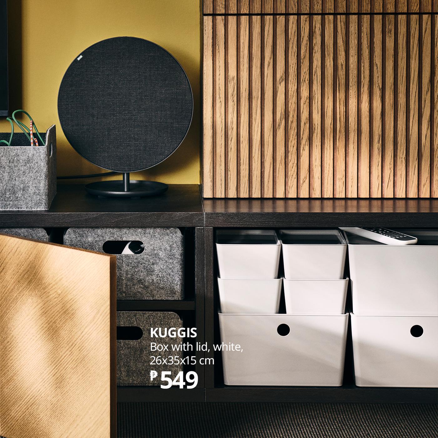A tidy IKEA shelf display features black and gray storage boxes, white KUGGIS bins, a round speaker, and warm wooden slats, creating a calm, modern feel.